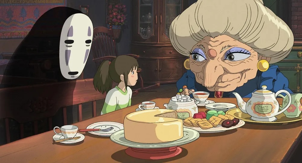 A still from the Studio Ghibli film Spirited Away showing Yubaba's tea party scene with a very english tea-looking spread of cake and tea and china crockery on the table with Yubaba, No Face and Chihiro seated at the table.