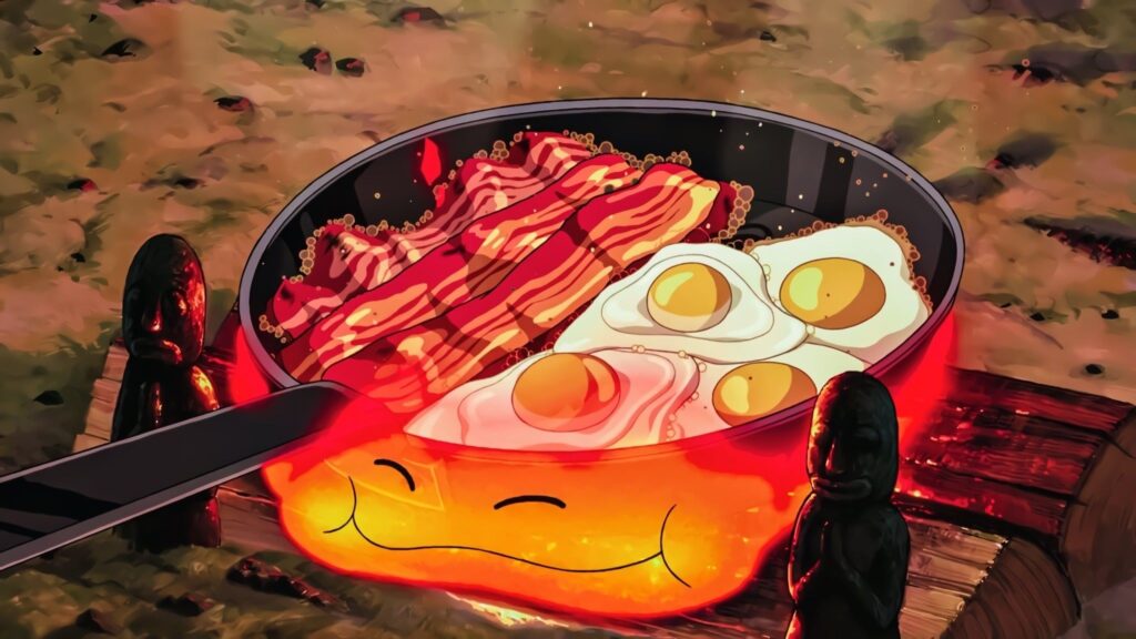 A still from Studio Ghibli's Howl's Moving Castle showing a breakfast of eggs and bacon frying in a pan over a flame, which has a smiling face.