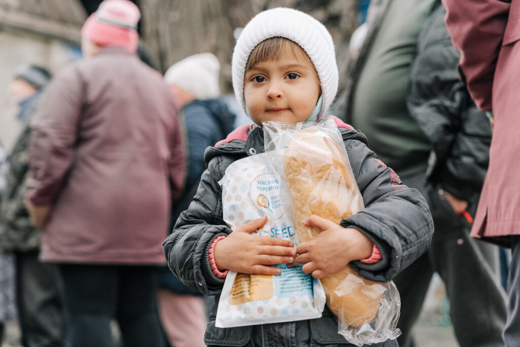 Portrait of a Ukrainian child in a woolly scarf holding a packet of seeds and a loaf of bread from World Central Kitchen