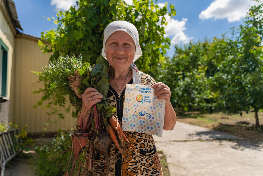Portrait of a Ukrainian woman in a headscarf holding an armful of freshly harvested vegetables and a packet of seeds from World Central Kitchen in the other hand