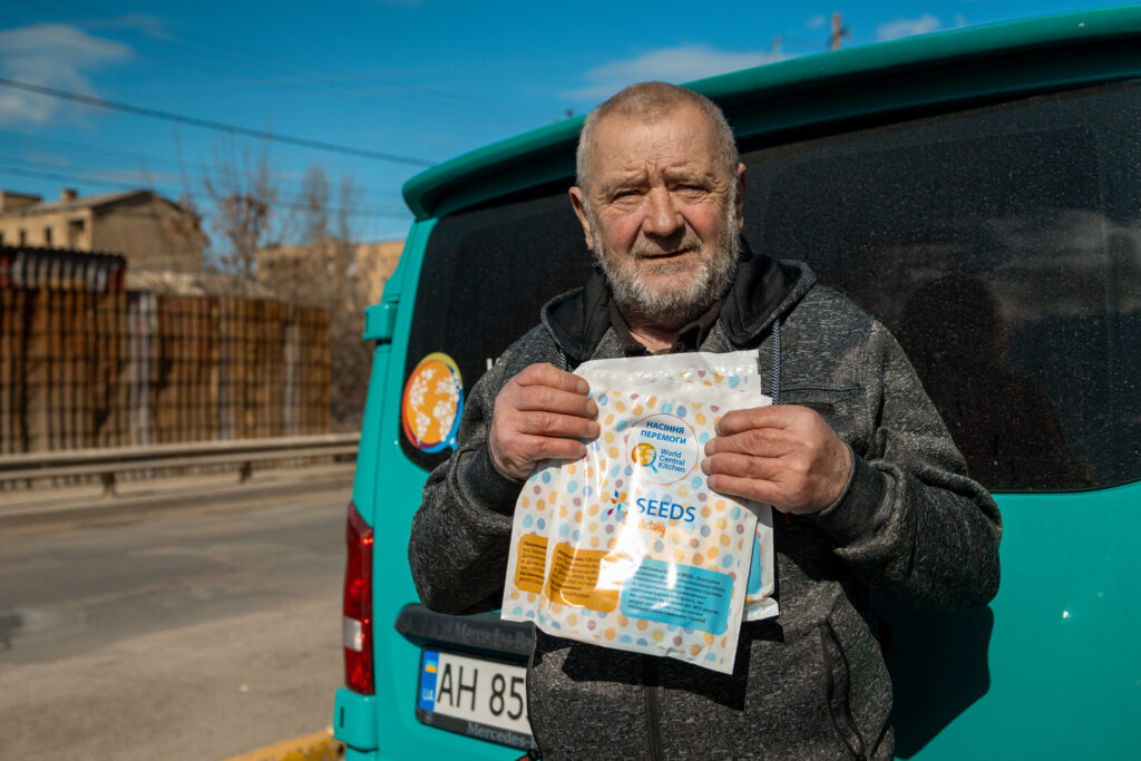 Portrait of a Ukrainian man standing behind a turquoise van and holding a packet of seeds from World Central Kitchen
